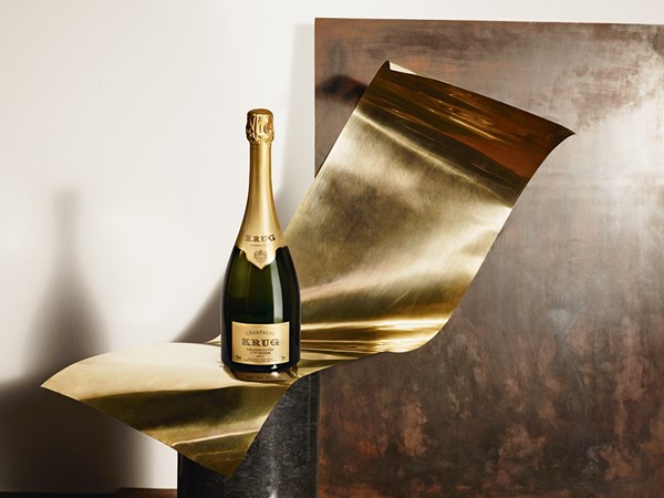 A bottle of Krug Grand Cuvée champagne elegantly rests on a curved sheet of golden metal, exuding a sense of luxury and refinement against a minimalist backdrop.