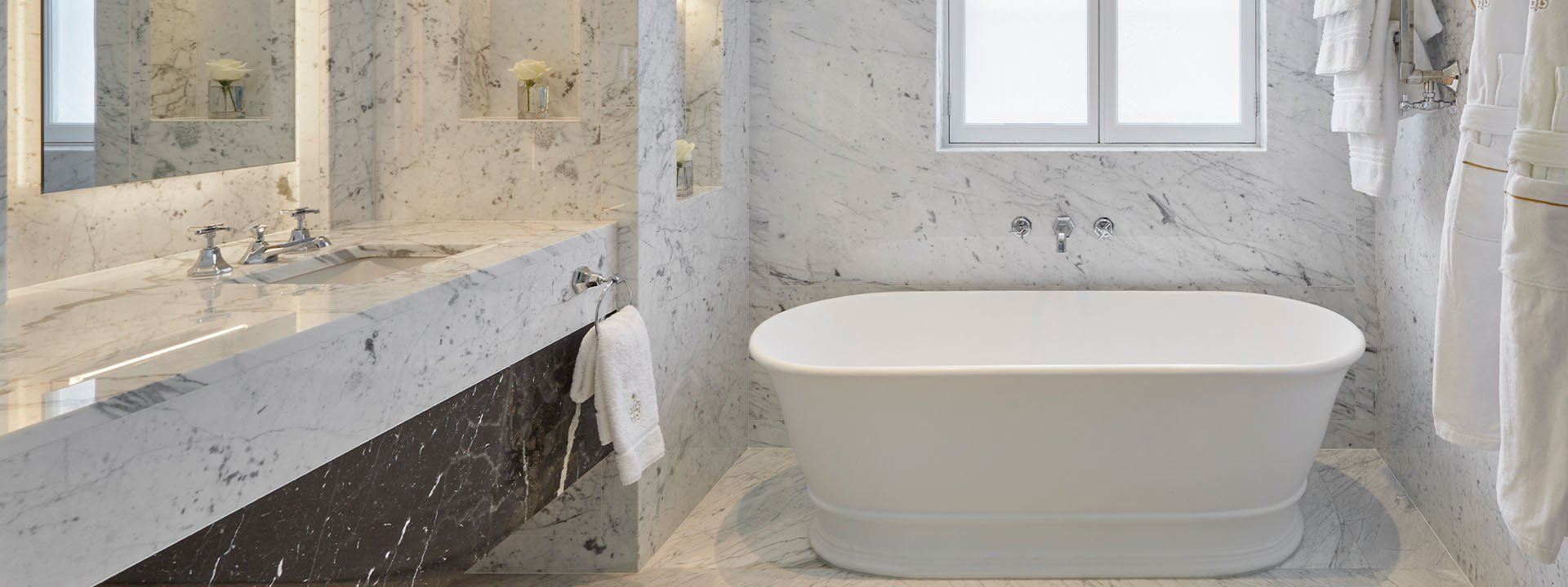 White marble bathroom with big freestanding bath tub at the end of the roo a sink to the left and dressing gowns and white towels hung up on the wall on the right. m