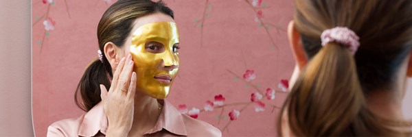 Woman looking at her reflection of herself in a gold MZ skin face mask. The background is pink.