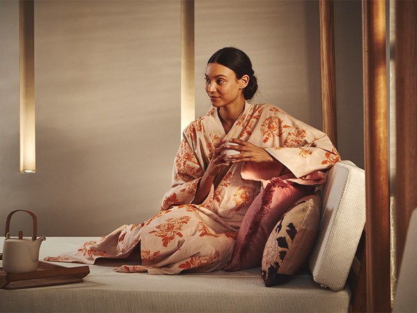 A woman wearing a floral robe sits peacefully on a cushioned daybed, holding a cup of tea, surrounded by soft pillows and warm ambient lighting, creating a serene spa-like atmosphere.