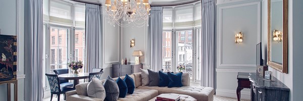 A luxurious suite with a curved wall of tall windows, elegant chandelier, plush sectional sofa, and soft blue decor accented by gold and marble elements.